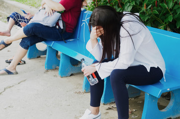 A girl in a white shirt with a bottle of drinking water in her hand, sitting on a bench holding her head in her hand