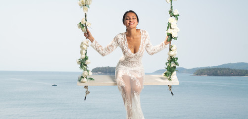 Beautiful woman in white lace dress posing on the outdoor swing with flowwers over sea and sky background