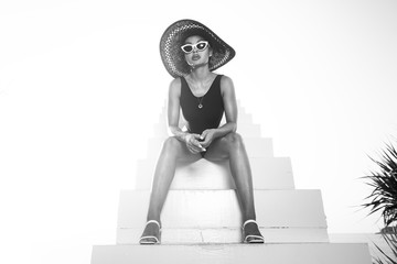 Wall Mural - Fashion girl in black swimsuit, hat and sunglasses posing outdoors on the stairs over sky background - black and white photography