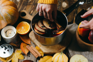 Woman's hand putting cinnamon stick in mulled wine hot with citrus, apple and spices in aluminum pot on wooden background. Autumn mood with pumpkins, candles and leaves.