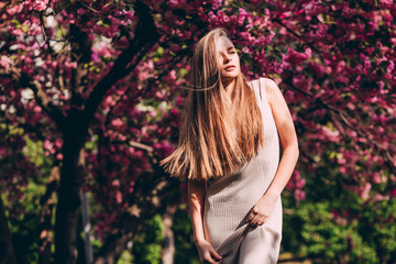 Wall Mural - Closeup portrait of a lovely girl in a park with blooming Japanese sakura trees. Romantic young blonde in a dress posing on a background of spring flowering trees. Cherry blossoms.