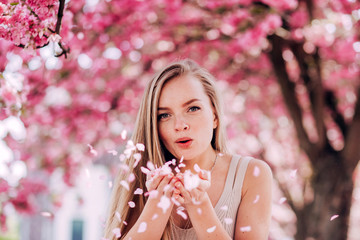 Wall Mural - Closeup portrait of a lovely girl in a park with blooming japanese sakura trees. Romantic young blonde in a dress posing on a background of spring flowering trees. Flying cherry petals.