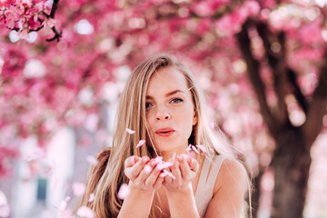 Wall Mural - Closeup portrait of a lovely girl in a park with blooming japanese sakura trees. Romantic young blonde in a dress posing on a background of spring flowering trees. Flying cherry petals.