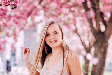 Wall Mural - Closeup portrait of a lovely girl in a park with blooming japanese sakura trees. Romantic young blonde in a dress posing on a background of spring flowering trees. Flying cherry petals.