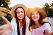 Two pretty girls take selfie in the summer outdoors at sunset. Girlfriends have fun, laugh, smile and take pictures. Closeup romantic portrait of two young women in dresses and straw hats.
