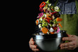 © Sergej - Chef throws a mix of vegetables in a metal bowl on a black background.