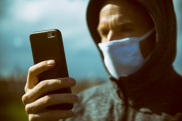 Serious man in a hoody wearing surgical mask checking his smartphone for coronavirus updates