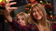 © Vit - Two pretty young girls or women are sitting near a New Year or Christmas tree and taking selfie on a mobile phone.