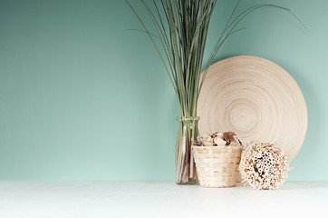 Calm eco home decor with natural beige dried plant, wicker basket, bamboo plate, bunch of reed in glass bottle, sheaf of twigs in green mint menthe interior on white wood desk.