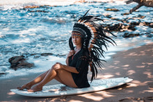 Beautiful And Confident Semi-nude Female Wearing American Indian Traditional Headdress, While Sitting On An Ocean Shore On A Surfer Board.