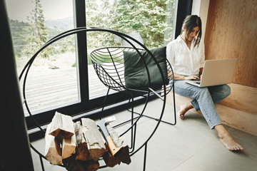 Stylish hipster girl with laptop sitting on floor at big window in modern room. Young woman using laptop, shopping or working online from home. Freelance and freelancer