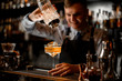 © fesenko - young smiling bartender pours drink from shaker into glass.
