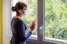 A woman under quarantine at home, wearing a medical face mask and a casual outfit, is standing idle in front of a closed window, the hand on the glass, staring into space.