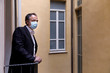 Uomo con mascherina chirurgica si affaccia al balcone con aria sconsolata, sullo sfondo la finestra di un edficio