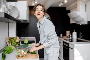 Wall Mural - Young and cheerful woman cooking food with healthy vegetarian ingredients on the kitchen at home. Healthy eating, food and lifestyle concept