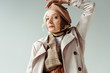 Fashionable elderly woman in trench coat looking at camera while posing isolated on grey