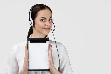 Beautiful smiling woman consultant of call center in headphones holding clipboard with blank white paper sheet on gray background. female customer support operator with headset