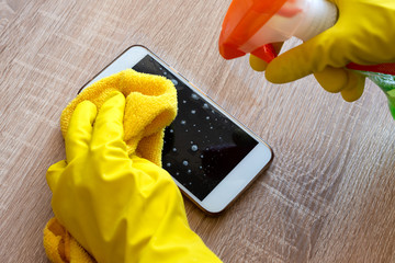 Female hand in rubber protective glove with rag wiping smartphone using cleaning spray.