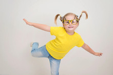 Happy Funny Little Girl In Party Bee Eyeglasses With Funny Pigtails Braids In Yellow T-shirt And Jeans.