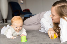 Cheerful Young New Mom Watching Baby Daughter Playing On Floor. Young Woman And Six Month Child Enjoying Leisure Time At Home. Motherhood Concept