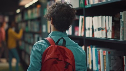 Wall Mural - University Library: Student Walks Between Rows of Bookshelves Searching for the Right Book Title for Class Assignment and Exam Preparations. Back View Following Camera Shot. Young People Study, Learn