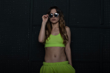 Wall Mural - Pretty cool young woman hipster in stylish sportswear straightens vintage sunglasses indoors. Attractive girl model in a trendy light green tracksuit posing near a vintage black wall in the room.