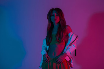 Wall Mural - Cool slim young woman in fashionable sportswear with a vintage hoodie posing with multicolored neon color background. Stylish attractive sexy girl in the studio with bright blue disco light.