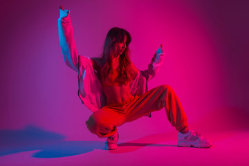 Wall Mural - Glamour young woman dancer in fashionable youth clothes in a white sneakers sits in a room with bright pink light. Modern girl model poses in the studio with neon amazing colorful red color.