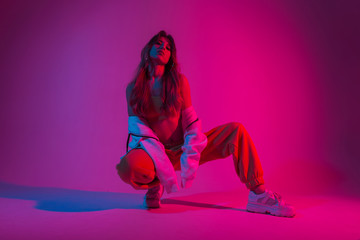 Wall Mural - Pretty attractive young woman dancer in fashionable youth clothes in a white sneakers sits in a room with bright blue light. Sporty beautiful girl model poses in the studio with neon colorful color.