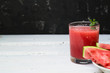 Slices of ripe watermelon on a plate and red watermelon juice in a glass on a wooden white background, and a black wall - side view. There is a place for text.