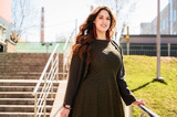Fototapeta Krajobraz - Photo of a pretty fashionable brunette woman with long beautiful hair with a smile in a suit on the background of the steps of the building in sunny weather