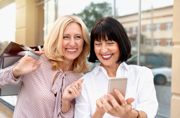 Wall Mural - Two mature women looking at the phone and having fun while doing shopping