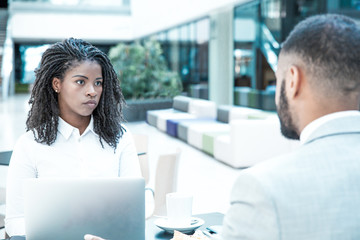 Diverse colleagues collaborating on project over cup of coffee in office lobby. Business man and woman sitting in cafe, using laptop, talking. Business breakfast concept