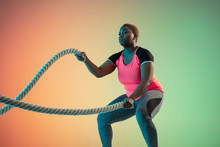 Young African-american Plus Size Female Model's Training On Gradient Background In Neon Light. Doing Workout Exercises With The Ropes. Concept Of Sport, Healthy Lifestyle, Body Positive, Equality.