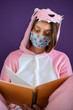 A girl in a medical mask and pink pajamas is reading a book, turning the page. Self-isolation in the house.Viral infection.