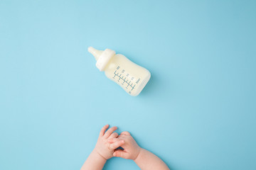 Infant hands and bottle of milk on light blue table background. Feeding time. Pastel color. Closeup. Point of view shot. Top down view.