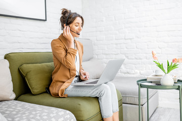 Young business woman working on computer, talking online using a headset while sitting on the comfortable sofa at home. Concept of remote work from home