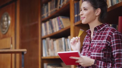 Wall Mural - Concentrated young woman standing in library and writing on notebook. University student completing assignment to submit while standing in library. Girl studying with copy space.