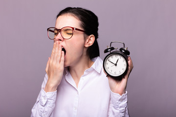 girl in a white shirt and glasses holds an alarm clock in her hand