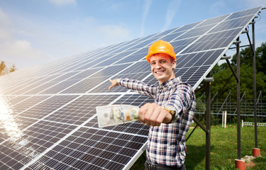 Wall Mural - Smiling guy with hundred dollar bill in hand shows at station of solar panels in which the sun's rays are reflected. Business and generation concept. Green ecological power energy generation.