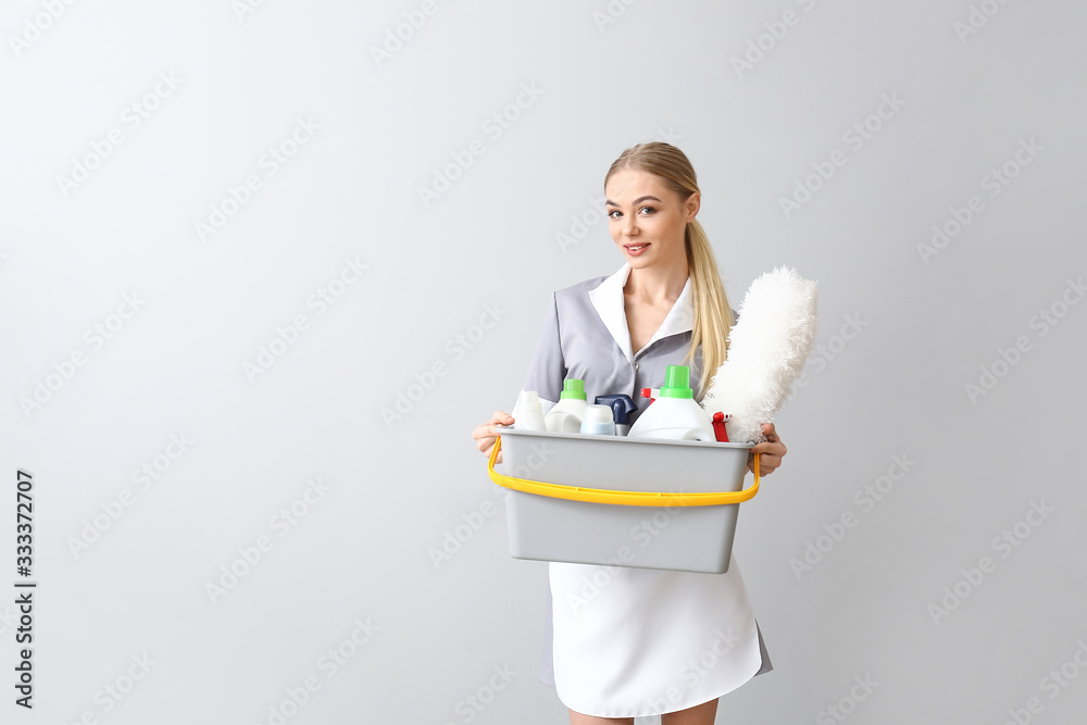 Beautiful young chambermaid with cleaning supplies on light background
