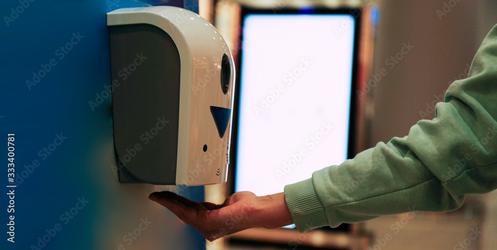 Macro shot of young woman using automatic hand sanitizer dispenser at the international airport with blank glowing advertisement banner on background. Close up, copy space. - obrazy, fototapety, plakaty 