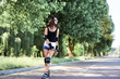 Young pretty brunette woman, riding rollerblades in city park with green trees. Fit sporty girl, wearing black top and white shorts, roller-skating in summer. Full-length portrait of slim sportswoman.