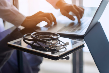 Male doctor hands typing on laptop computer keyboard, search medical information with digital tablet pc and medical stethoscope on the desk at office. Online medical,medic tech, emr, ehr concept.  