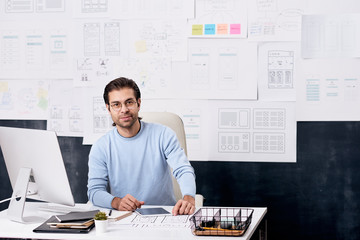 Wall Mural - Portrait of content handsome UI designer in eyeglasses sitting at desk with computer in modern office with task board