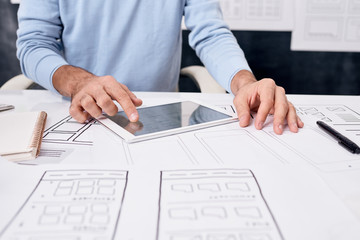 Wall Mural - Close-up of unrecognizable man sitting at table with sketches and using tablet while developing app design