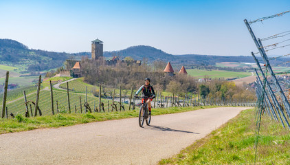 Wall Mural - ageless happy senior woman riding her elechtric mountain bike on sunny springtime day in the Vineyards of the Bottwartal with beautiful medieval castle in the background, Heilbronn, Baden-Wuerttemberg