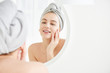 © amixstudio - Portrait of  young girl with  towel on head in white bathroom looks and touches her face in the mirror and enjoys youth and hydration. Natural beauty, home care for problem skin