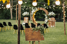 Handmade Wooden Board With For Welcome Sign Decorated With Green Plants.  Board Welcome To Our Wedding  On Reception In Garden With Wooden Arch And Black Chairs On Background. Copy Space. 