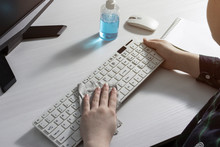 A Girl In A Checked Shirt Cleans The Keyboard With An Antibacterial Rag. View Over The Girl's Shoulder. Cleaning Agent, Mouse And Notepad On A White Office Desk.